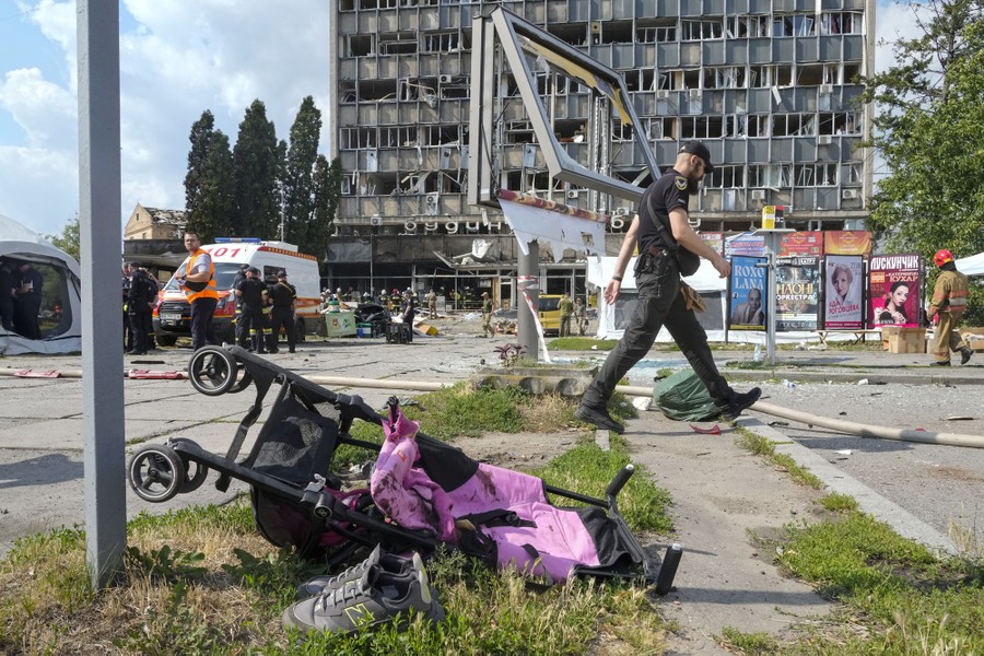 First responders stand and walk near a missile-damaged building, with a toppled baby stroller seen in the foreground.