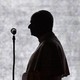 Photograph of Pope Leo’s black silhouette from the side as he speaks in front of a microphone.