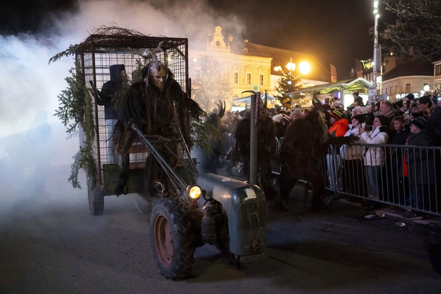 A person wearing a Krampus costume drives a tractor that carries two people in a cage, in a parade.