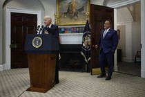 Joe Biden gestures at a White House podium, Miguel Cardona stands by the open door