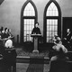A black-and-white photo of Charlie Chaplin at a podium, surrounded by onlookers in pews