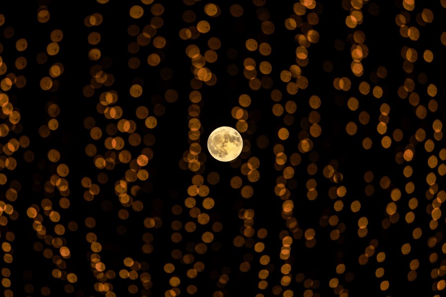 The full moon rises, seen through a cluster of small, blurry lights.