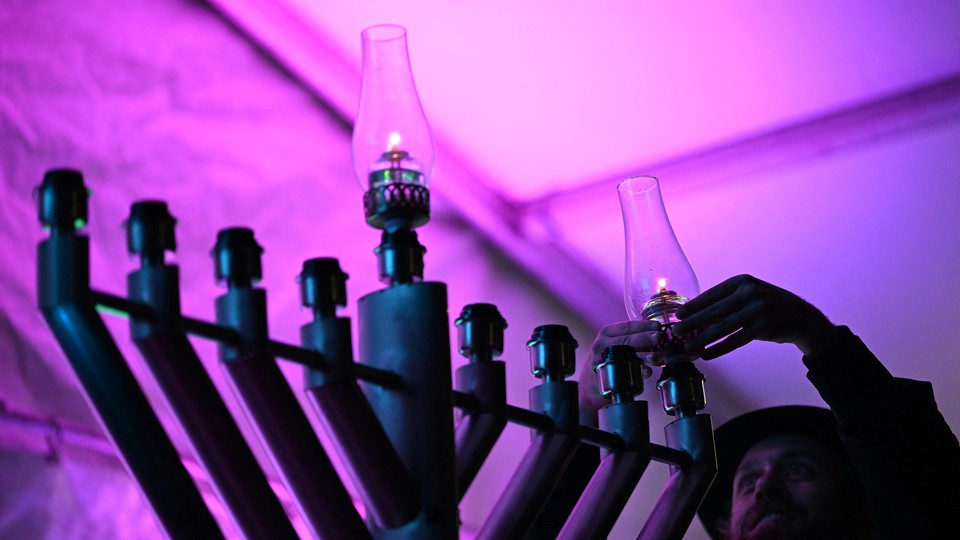Photograph of a man lighting candles on a menorah in a purple-lit room