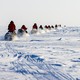 About a dozen people in red parkas ride snowmobiles in a line across a broad, flat expanse of snow and ice.