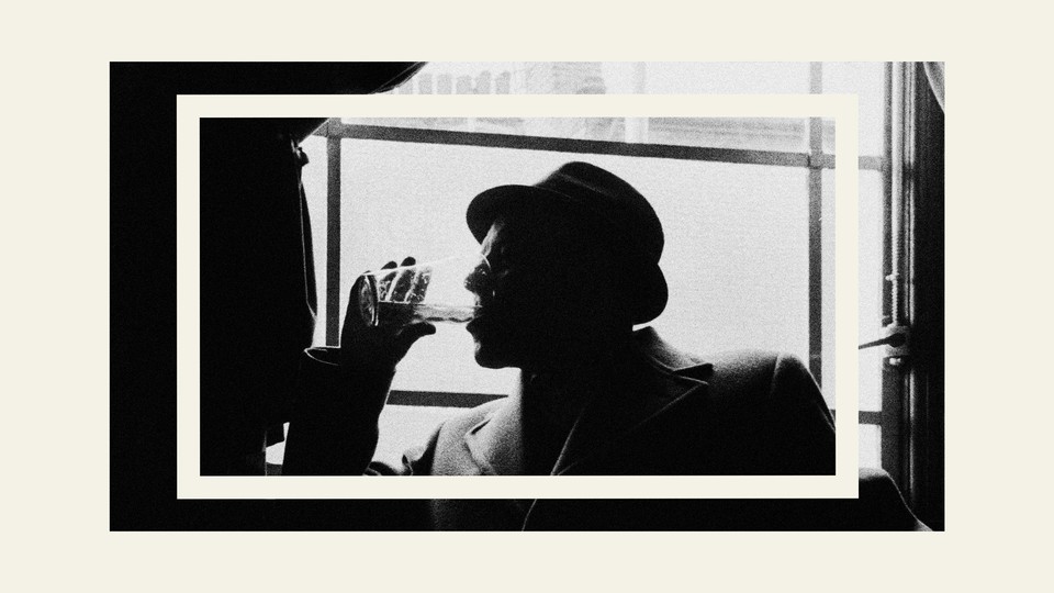A black-and-white image of a man in side-profile wearing a hat and a coat, drinking from a clear glass