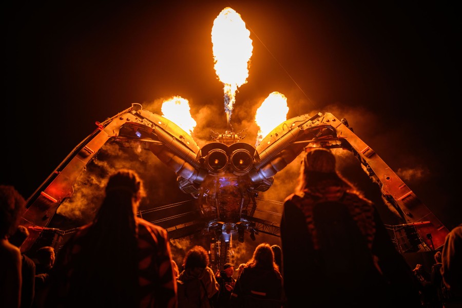 Fire erupts from the top of a large robotic spider sculpture as people look on below.