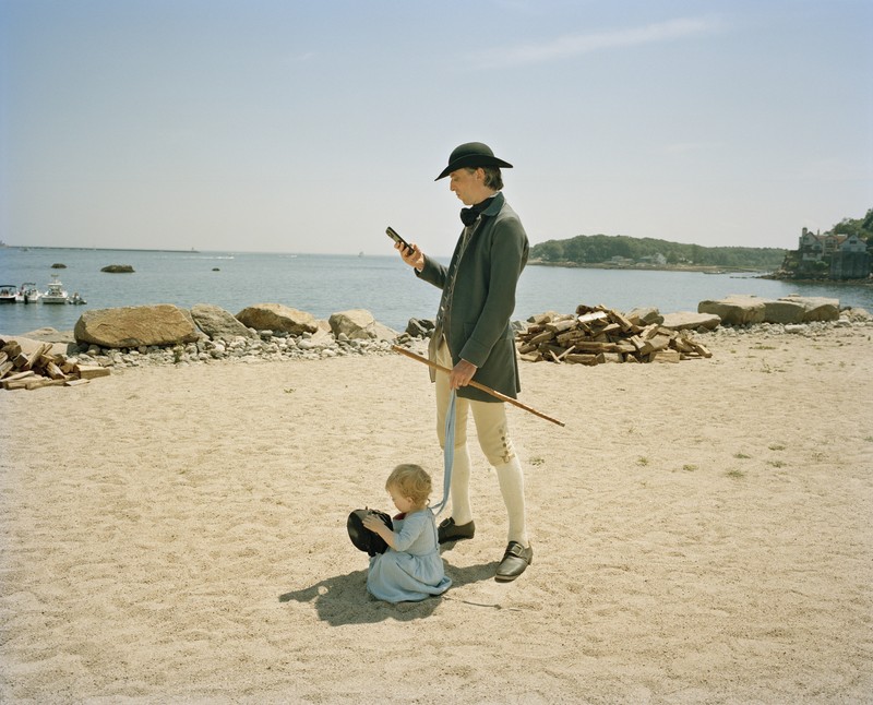 photo of male reenactor in period dress with hat, coat, breeches, and cane standing on sandy beach looking at his cell phone, with very young child playing at his feet