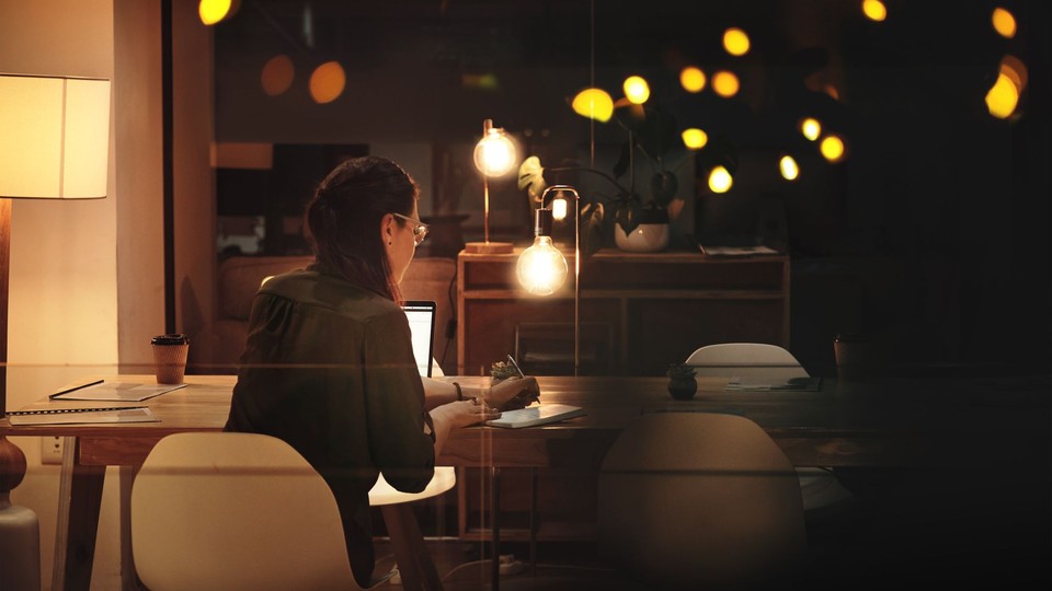 dark-haired woman sitting at a table writing in a notebook, back to the camera, lights reflected in the background