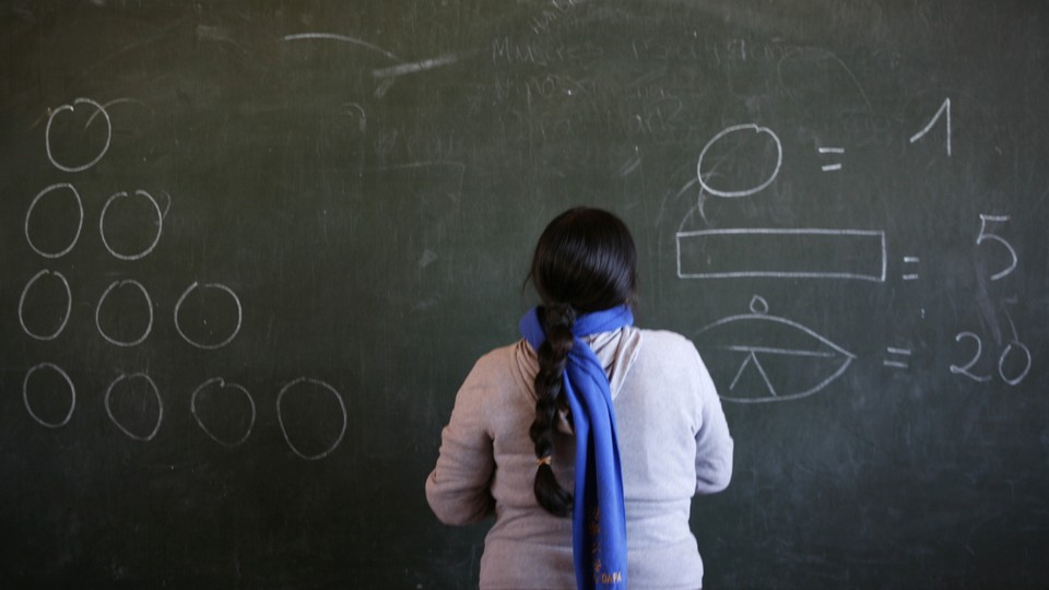 A teacher faces a chalkboard with numbers and shapes on it.