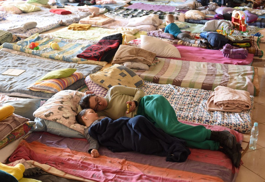 A woman and a child lie side-by-side on a foam mat and pillow, among many such makeshift beds, on the floor of a large room.