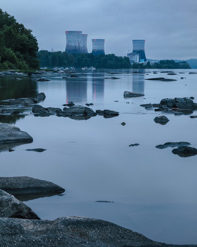 photo at dusk of river with rocks protruding and island in distance with 4 huge cooling towers with red lights
