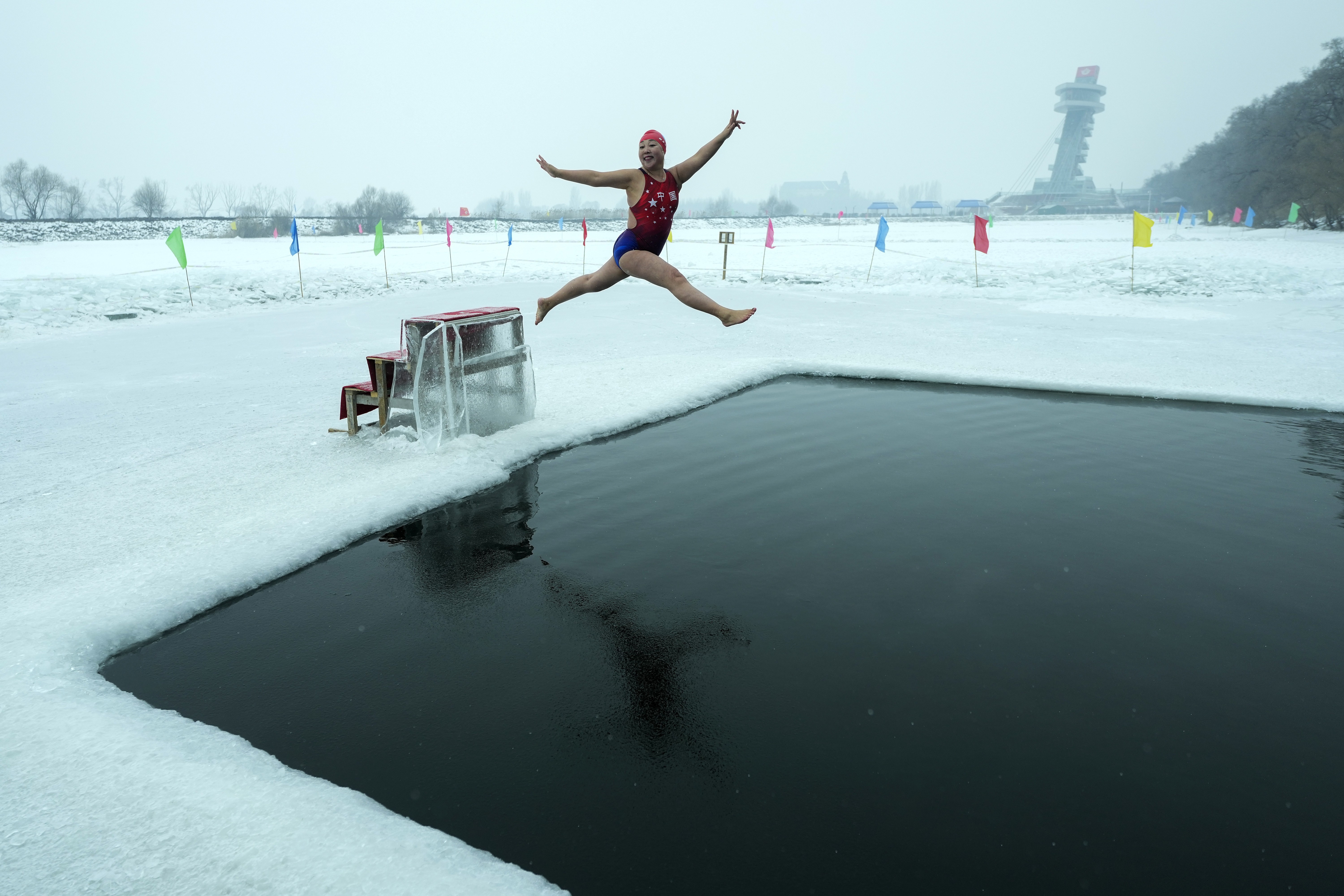 A woman in a bathing suit leaps from a small platform, about to plunge into freezing water in a pool cut into a frozen river.