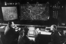 A black-and-white photograph of a group of people looking at a screen displaying a map of the United States