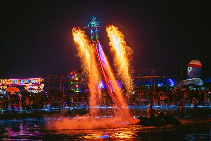 A performer stands on a platform lifted in the air by water jets, while holding a long pole with iron baskets on either end that throw off plumes of sparks.