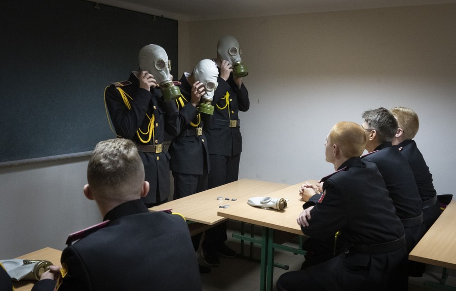 Three military cadets standing in front of a classroom put on full-head gas masks.