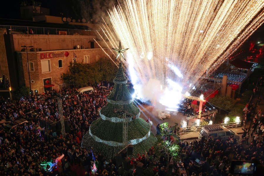 A large crowd watches fireworks erupt near a Christmas tree in a city square.