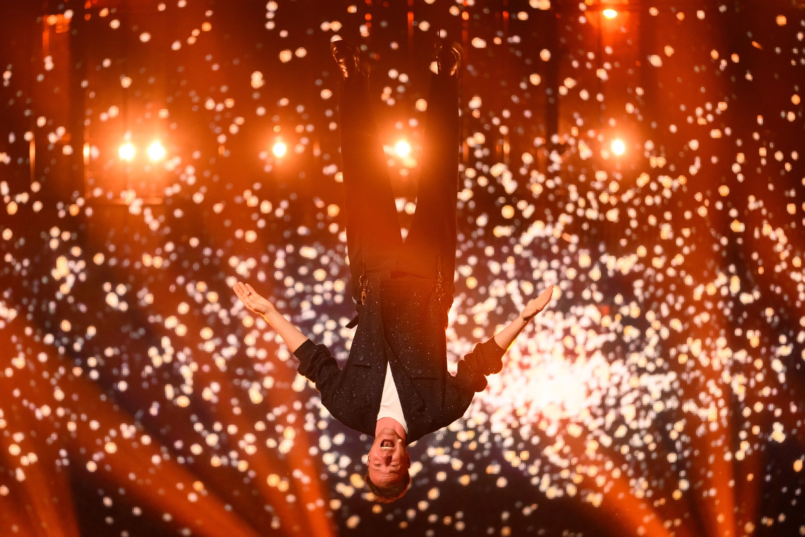A singer performs while being suspended upside-down over a stage, with sparks projected in the background.