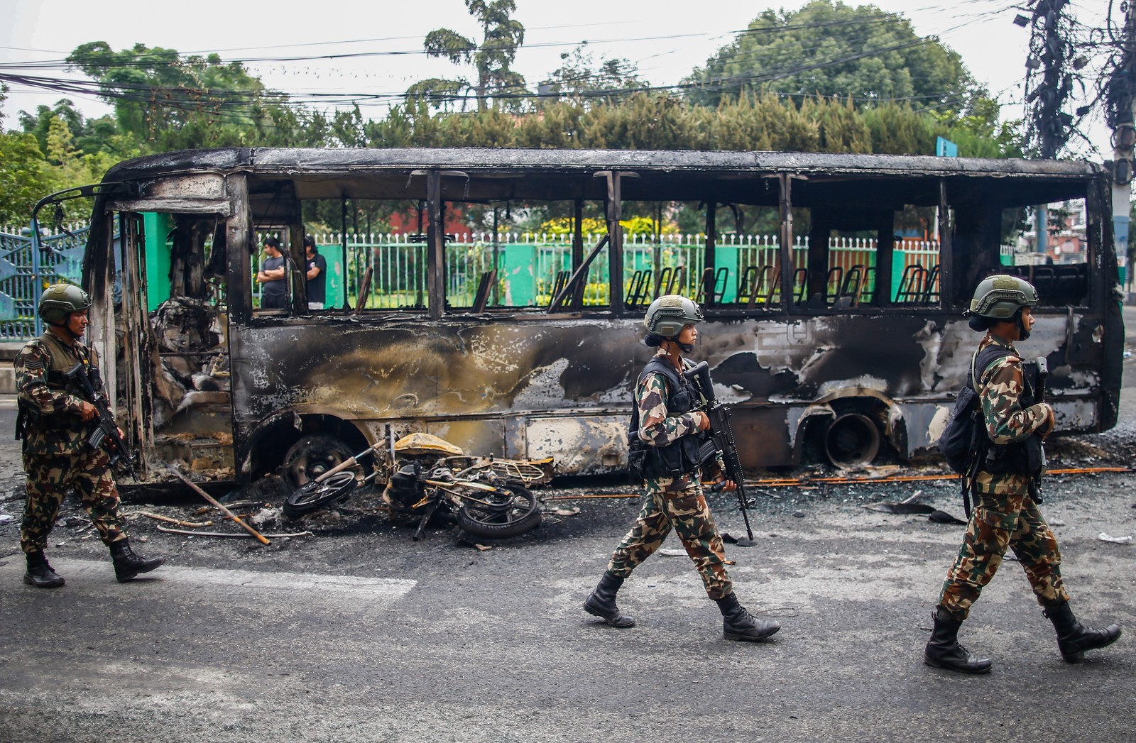 Several soldiers walk past a burnt-out bus.