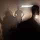 Photograph of black silhouettes of border agents standing with hands on their hips illuminated by a vehicle’s headlights behind them