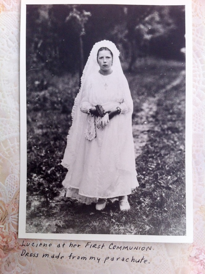 Photo of a black and white photo in an album, of a young girl wearing a white dress. Handwriting under the photo says: "Luciene [sic] at her First Communion. Dress made from my parachute."