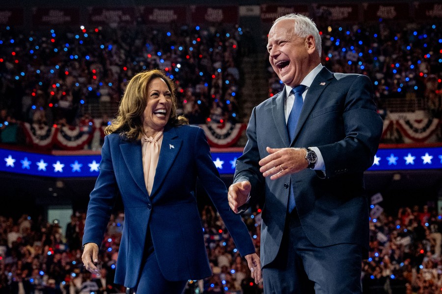 Vice President Kamala Harris and Minnesota Governor Tim Walz laugh as they walk together on a stage, in front of a crowd.