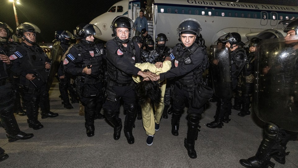 A prisoner with his head pushed down is escorted from a plane by a large group of security forces