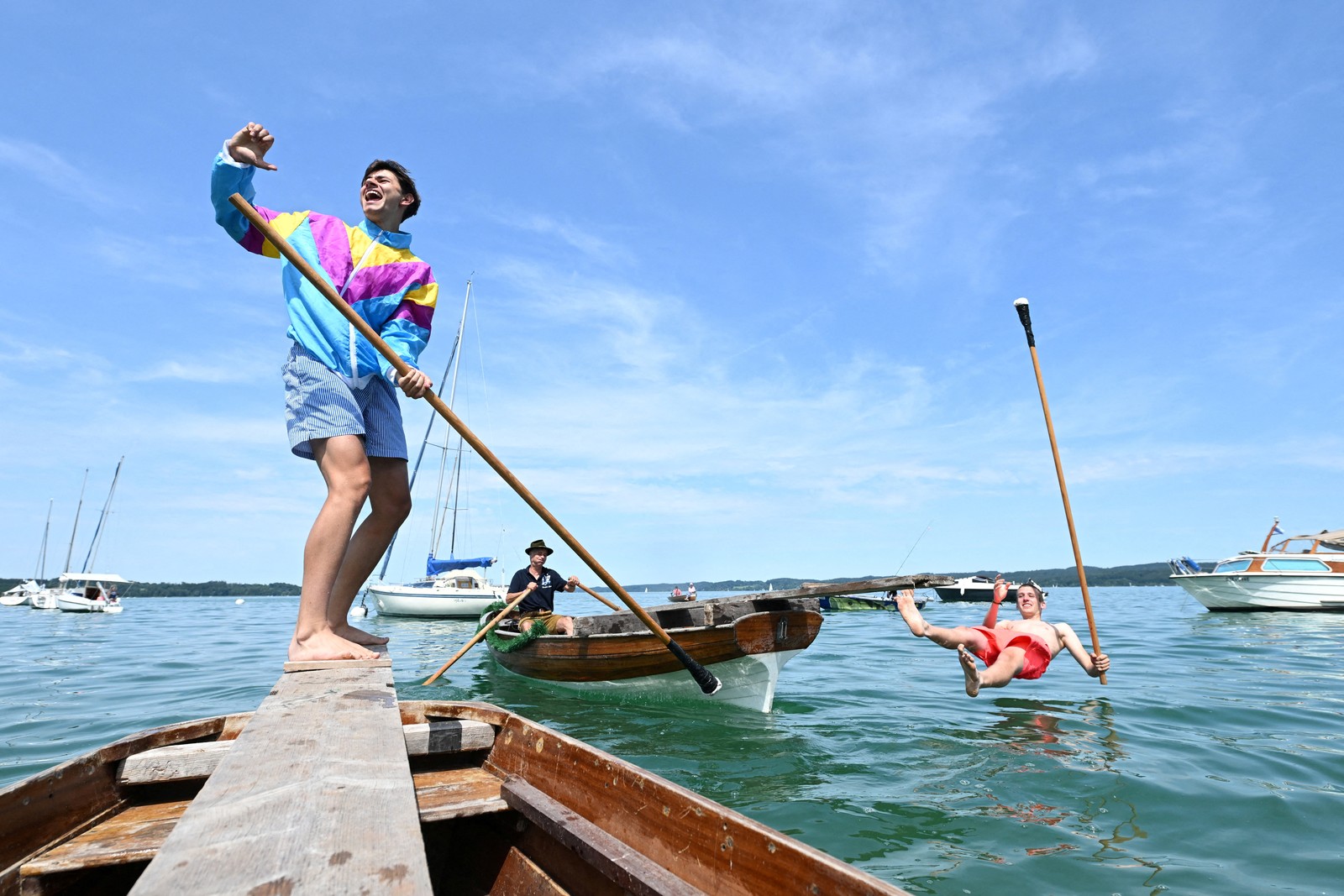 One person stands on a small boat, as another falls into the water after a jousting match.