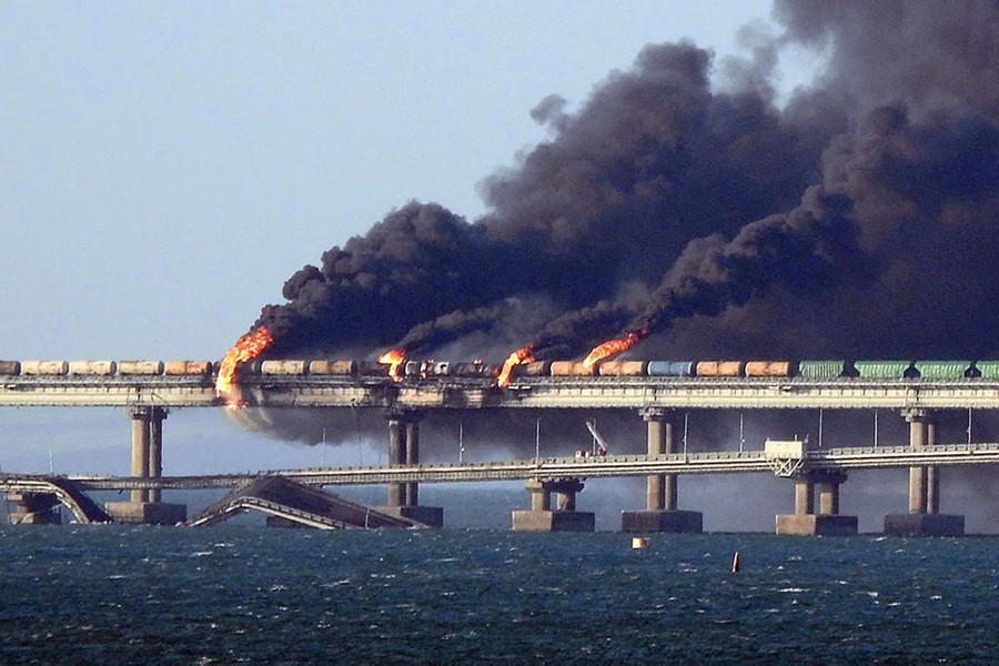 Multiple fires burn along a cargo train on a long bridge over water, beside a parallel road bridge that has been partially destroyed.
