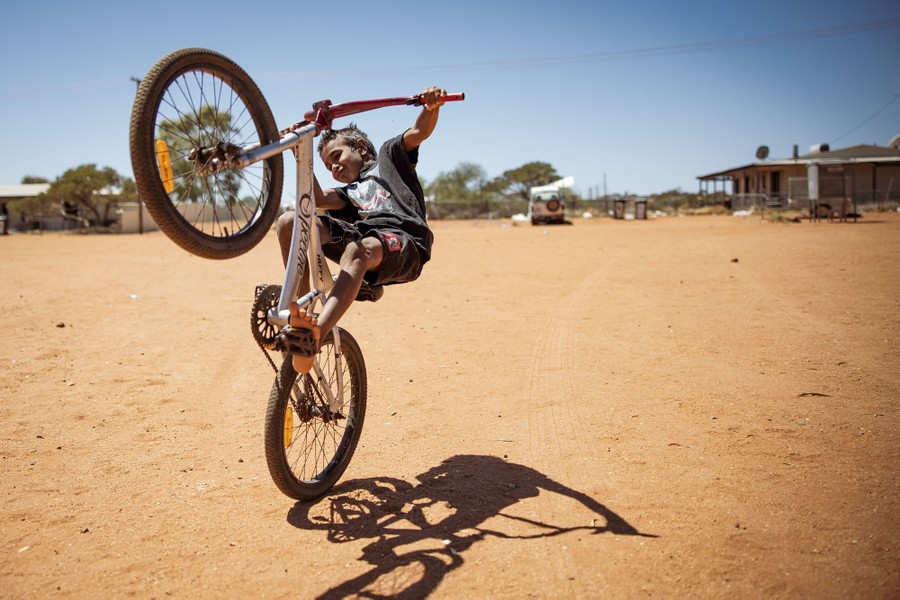 A child pops a wheelie on a bike on a patch of dirt near some houses.