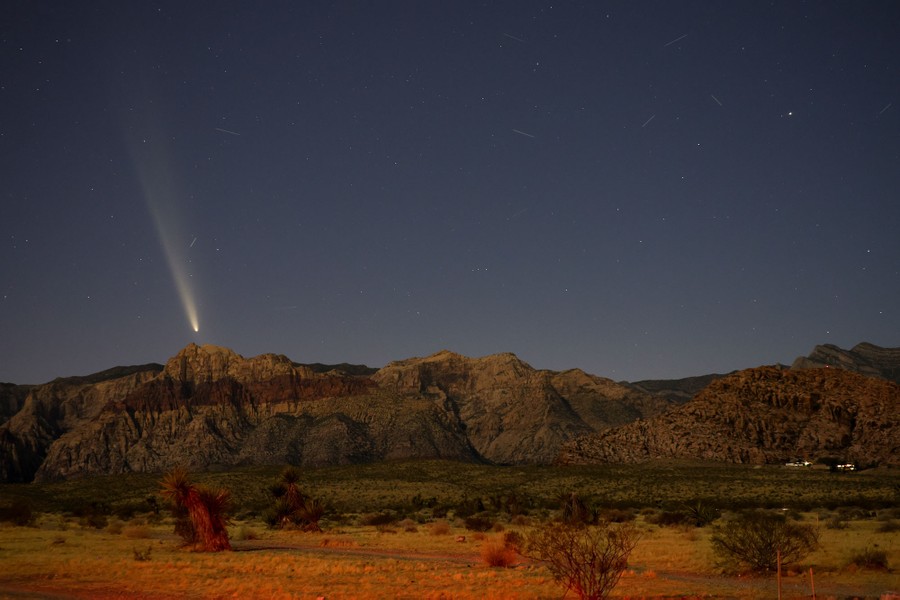 A comet appears in the night sky above the horizon in a desert.