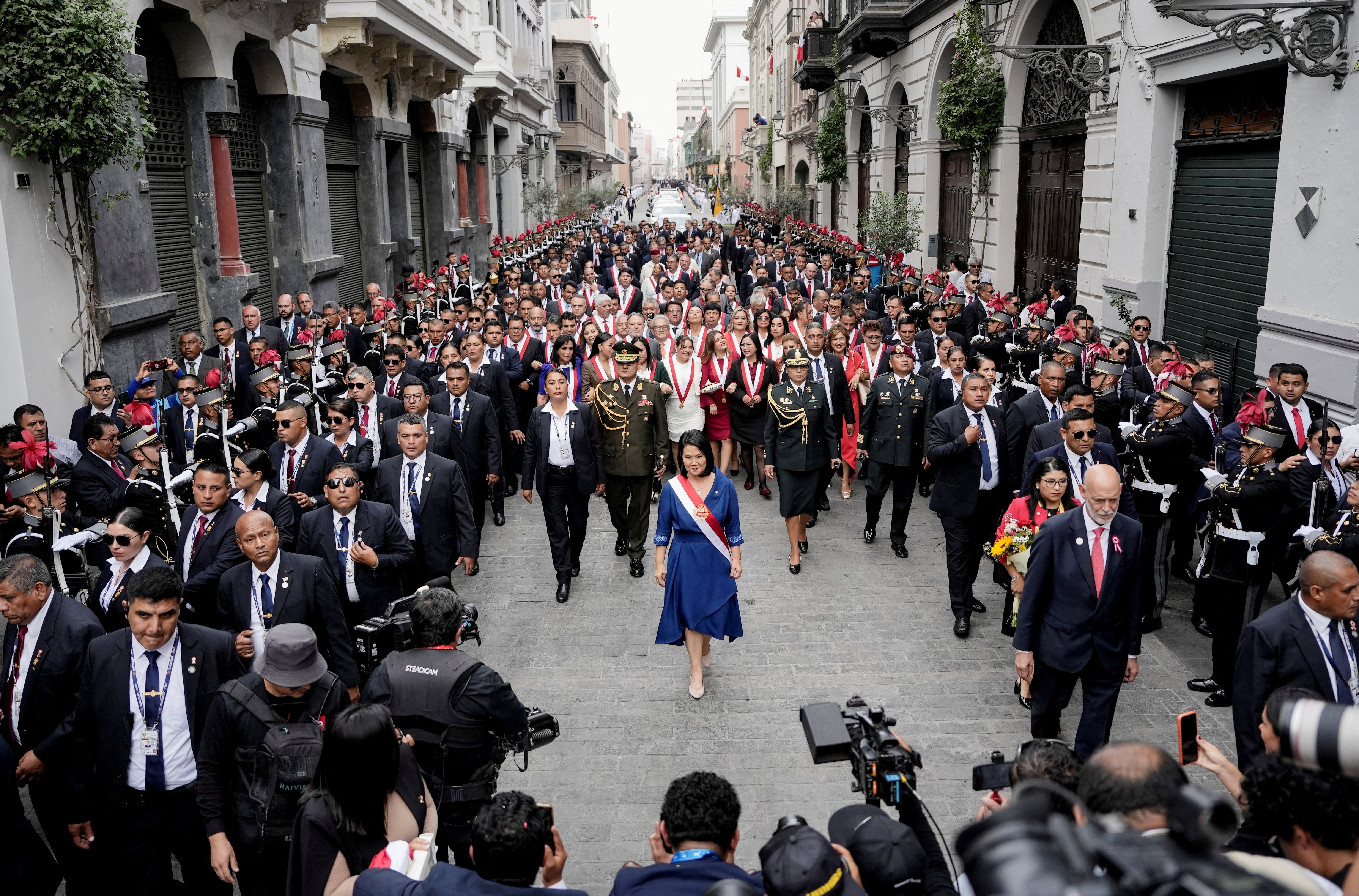 Peru’s President Keiko Fujimori walks down a cobblestone street, surrounded by soldiers, bodyguards, and other government officials.
