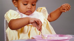 Color photo of a small child wearing a bib and eating off a pink plastic plate using a spoon
