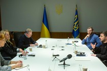 photo of large white meeting table with two women and a man on left and two men on right, one speaking, with Ukrainian flags in background