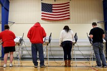 The backs of voters as they cast their ballots. They stand underneath an American flag, presumably in a gymnasium.