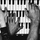 Black-and-white photo of many different hands playing three stacked sets of piano keys.