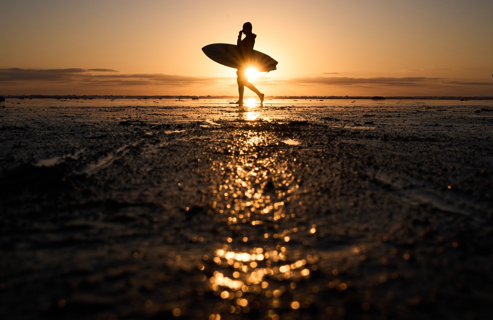A surfer carries their board, walking on a beach at sunset.