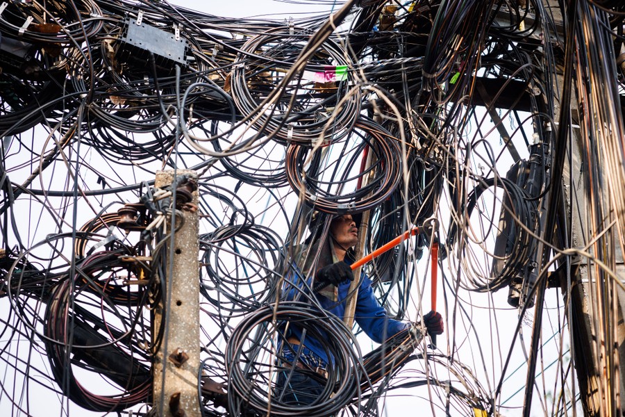 A worker uses wire cutters while under a massive tangle of cords and wires hanging from poles.
