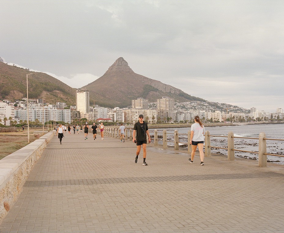 photo of paved promenade along curving shoreline with people walking and excercising, with city buildings and large, jutting mountain ridge in background 