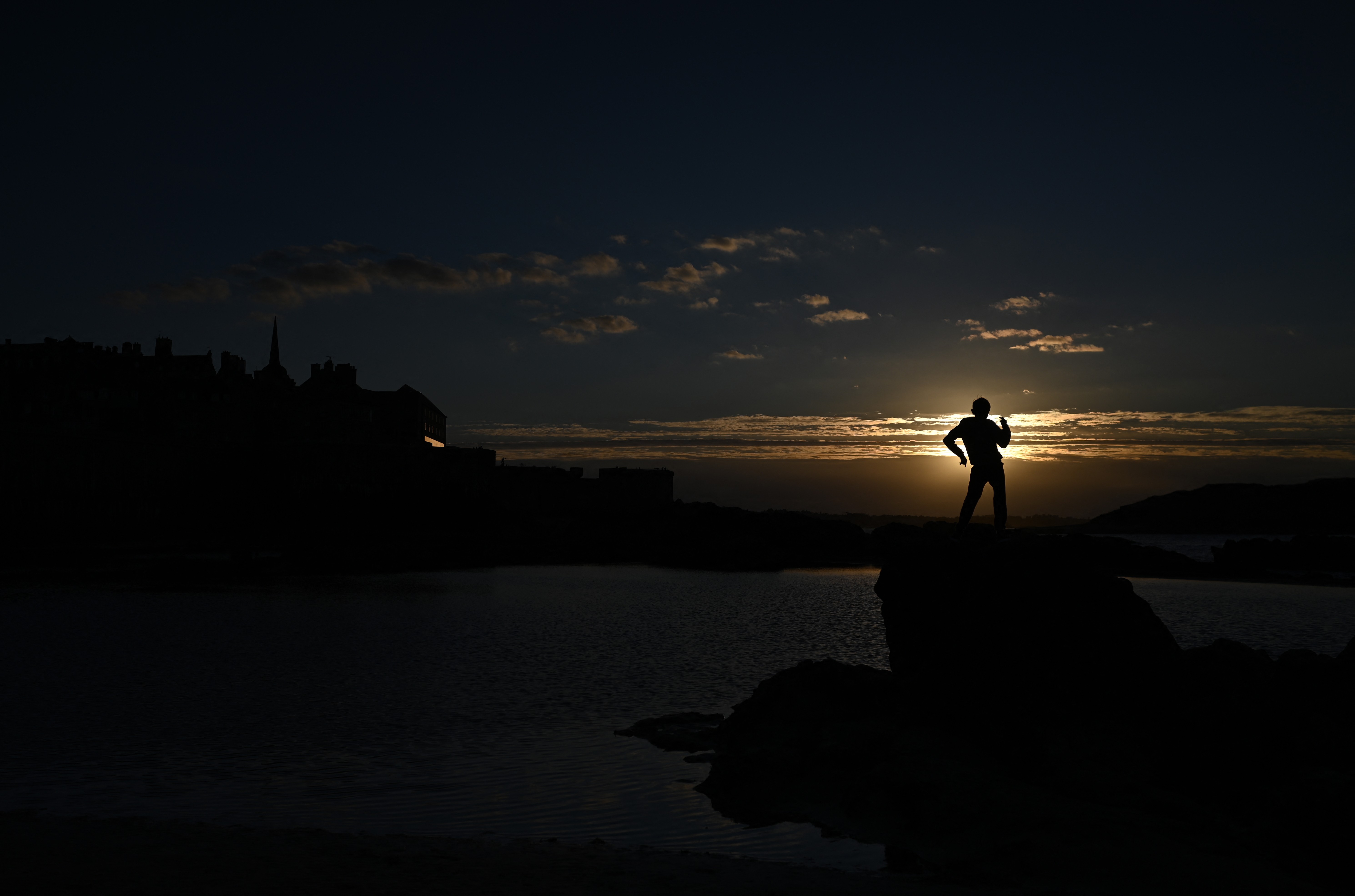 A boy plays on rocks on a beach, silhouetted by the setting sun.