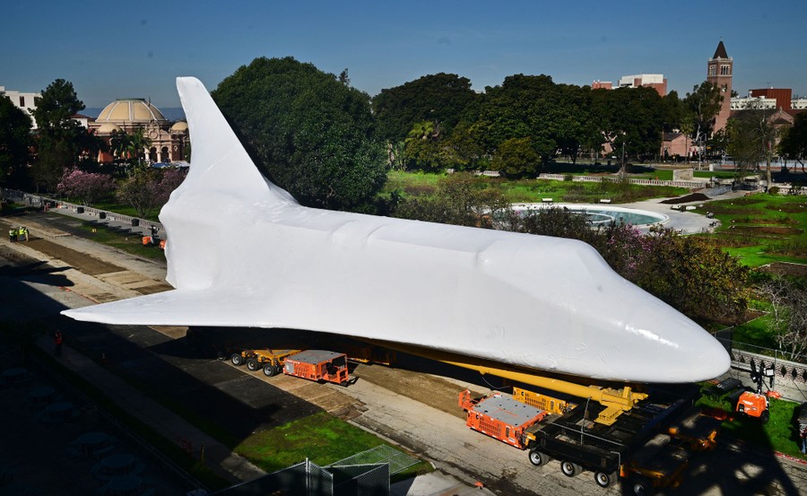 A space shuttle, wrapped in white plastic, is carried on a trailer through a park.