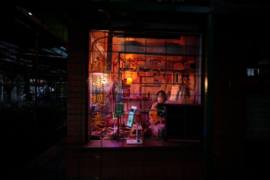 A worker sits inside a small food store behind glass and barriers.