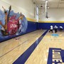 A group of children plays basketball on a brand new gymnasium floor. A mural with musical instruments and notes is pained with the school's name on the wall.