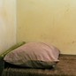 A twin bed in the corner of a room, with a thin brown blanket and a pink pillowcase, against a stained wall