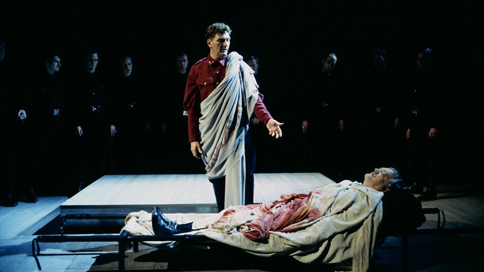 A bloody actor lies on a cot while another actor speaks and others look on.