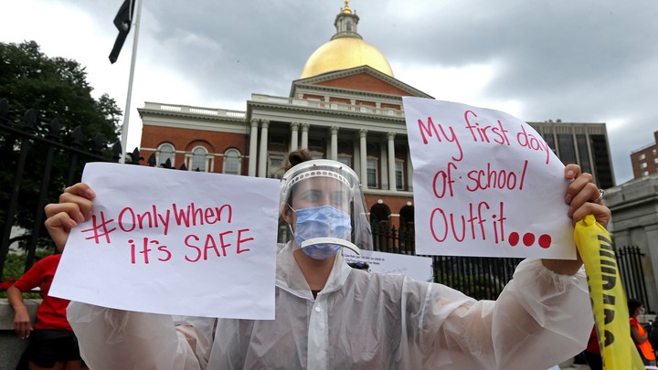 A woman wearing a face shield, surgical mask, and plastic poncho holds up signs in protest of school reopening.