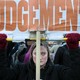A photo of a protester holding a sign that reads "Judgement" in orange letters on a white background