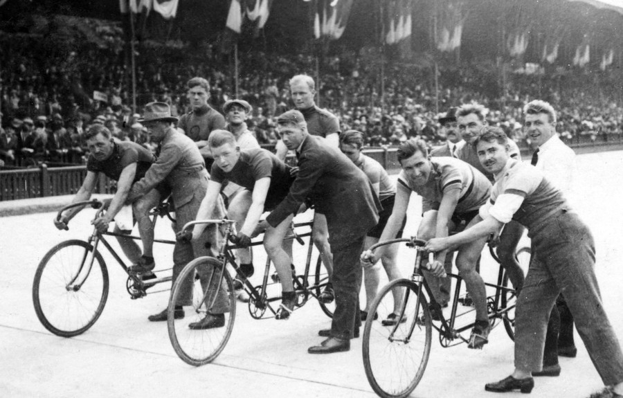 People help hold up three tandem bicycles and riders on a track as they prepare for a race.