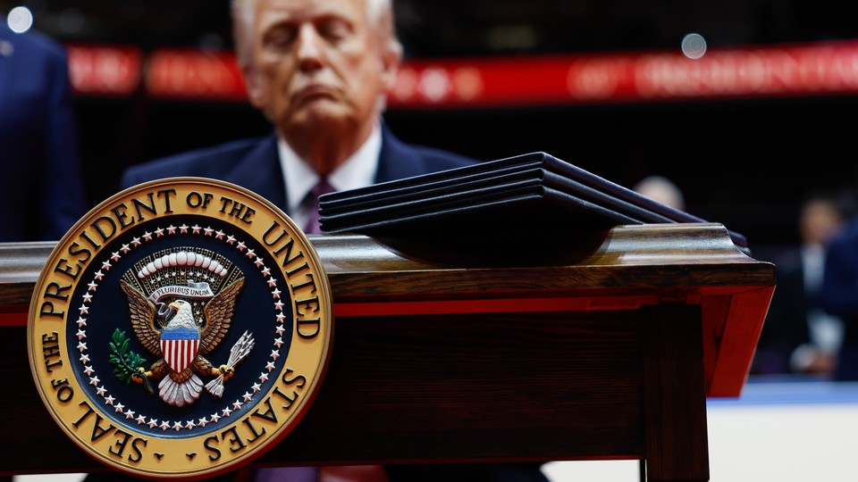 Photograph of Donald Trump behind a desk with the presidential seal and executive orders atop the desk