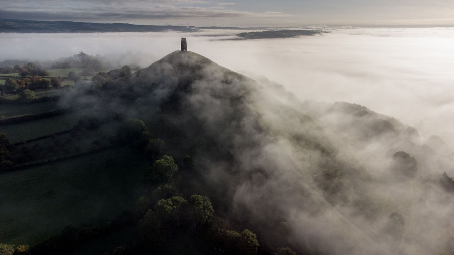 An aerial view of mist covering fields, leading up to a hill topped by a stone tower.