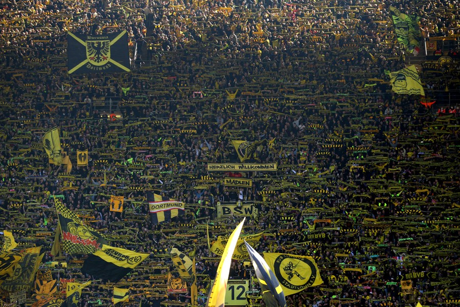 A large crowd fills a stadium, waving black-and-yellow banners before a football game in Germany.